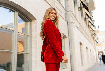 Beautiful smiling woman  in  elegant red velvet suit holding purse and  posin outdoor in old european city.    Blond wavy hairs, perfet skin, full lips.