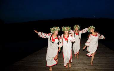 Happy girls in embroidered dresses and wreaths on their heads dance on the pier against the...