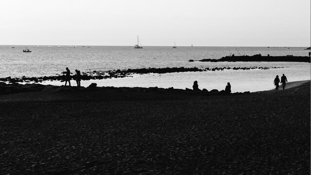 B&W seascape with all what you can see in a beach overview with people silhouettes relaxing at sunset, they speaking,  walking, fishing and did outdoor activity with sail boats