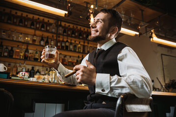 A bearded man in an elegant suit raises a glass of whiskey in a bar.  A man toasting a glass with an expensive alcoholic drink near bar counter