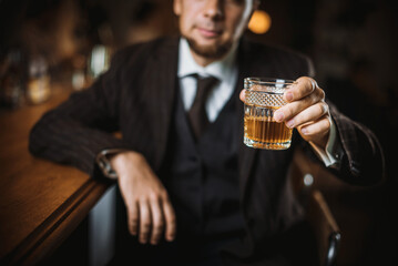 A bearded man in an elegant suit raises a glass of whiskey on a dark background. Toast at the bar....