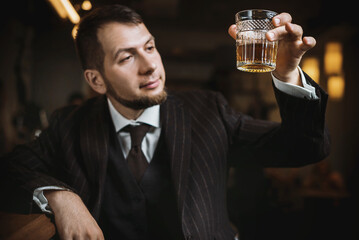 A bearded man in an elegant suit raises a glass of whiskey in a bar.  A man toasting a glass with an expensive alcoholic drink near bar counter