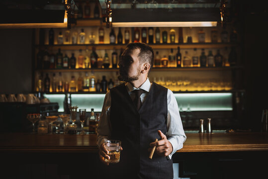 A bearded man in an elegant suit standing near bar counter in a dark. A man toasting a glass with an expensive alcoholic drink. Night atmosphere in luxury restaurant