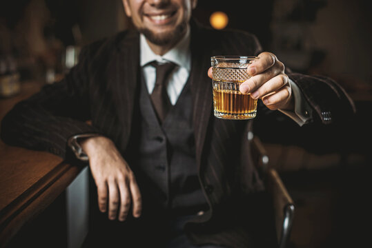 A bearded man in an elegant suit raises a glass of whiskey on a dark background. Toast at the bar. An unrecognizable man holds a glass with an expensive alcoholic drink on a blurred background. select