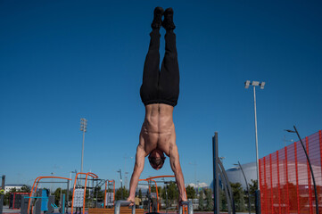 Shirtless man doing handstand on parallel bars at sports ground. 