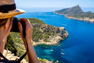 A female photographer with hat shots a picture from a cliff of the rocky coastline of idyllic...