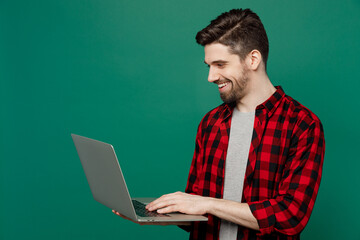 Young happy smiling cheerful fun cool man he 20s wearing red shirt grey t-shirt hold use work on laptop pc computer isolated on plain dark green background studio portrait. People lifestyle concept.