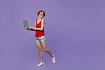 Full body side view young fun smiling woman 20s she wear red tank shirt eyeglasses hold use work on laptop pc computer look aside on workspace area mock up isolated on plain purple backround studio