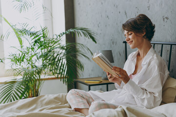 Side view young calm happy woman wear white shirt pajama she lying in bed read book study rest...