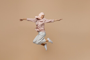 Full body young fun arabian asian muslim woman she wear abaya hijab pink clothes jump high with outstretched hands isolated on plain pastel light beige background. People uae islam religious concept.