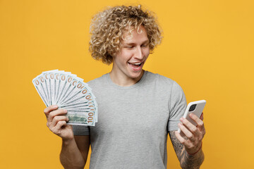 Fototapeta Young smiling happy caucasian man 20s he wear grey t-shirt holding fan of cash money in dollar banknotes mobile cell phone isolated on plain yellow backround studio portrait. People lifestyle concept. obraz