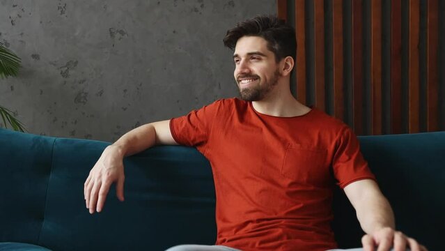 Young smiling happy satisfied man wears casual clothes red t-shirt sits on blue sofa couch stay at home hotel flat rest relax spend free time in living room indoors grey wall. People lounge concept
