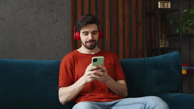 Vivid young man wears red t-shirt listen music in headphones use mobile cell phone dance gesticulate hands sits on sofa stay home spend free time in living room indoors grey wall People lounge concept
