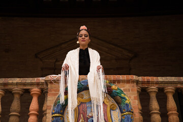A beautiful teenage flamenco dancer with brown hair stands on a balcony wearing a shawl embroidered...