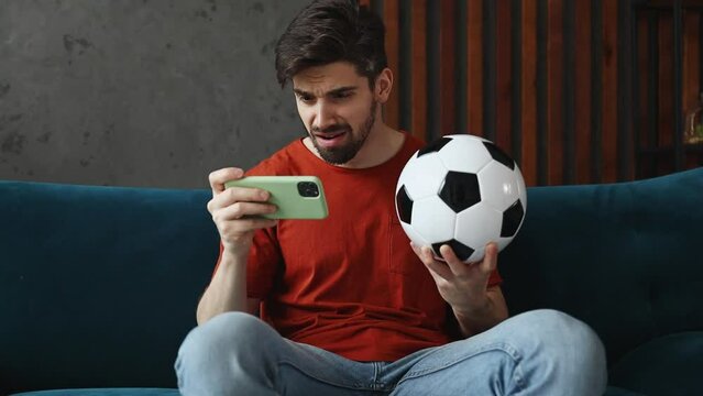 Young man wears red t-shirt support football team watch tv live stream use mobile cell phone do winner gesture sits on blue sofa stay home spend free time in living room indoors People lounge concept