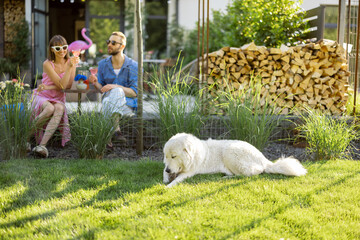 Young stylish couple have a drink while sitting together with dog on a lawn at their beautiful...