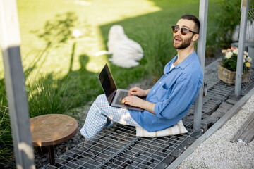 Cool guy works on laptop while sitting at green lawn of country house backyard. Concept of remote...