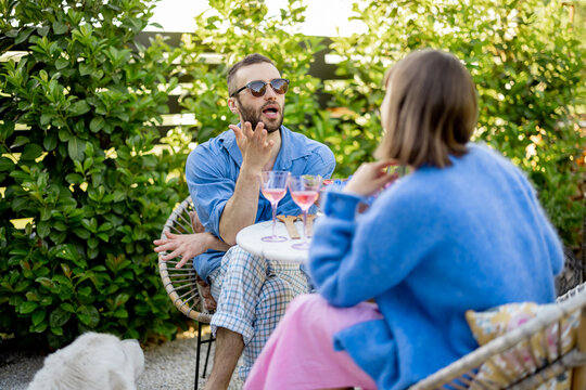 Young stylish couple have conversation while sitting by the round coffee table at their beautiful garden. Young man and woman have romantic evening outdoors - Powered by Adobe