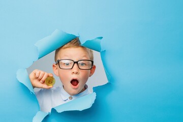 Happy cute businessboy in eyeglasses is having fun and holding bitcoin in hand on blue background wall, climbs through a hole in the paper. Cryptocurrency and modern finance.