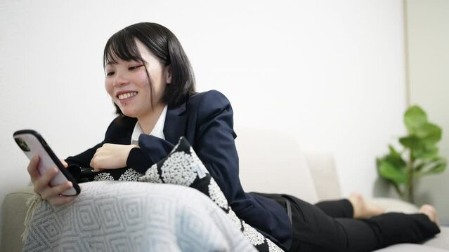 リビングのソファでスマートフォンを操作するスーツ姿の可愛い日本人女性 Cute Japanese woman in a suit operating a smartphone on a sofa in the living room. 