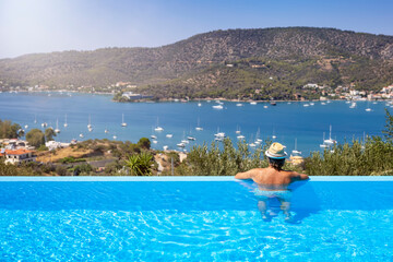 A tourist man in a swimming pool enjoys the view of the sailing boats in a bay at Poros island,...