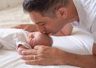 Fototapeta na wymiar Happy father kisses his newborn. Baby sleeping soundly and peacefully. Man in white t-shirt with happy smile. New worries and style life. Concept of child care, feeling safe, parent love