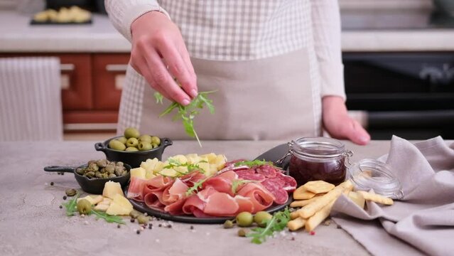 making meat and cheese antipasto plater - woman pouring arugula to serving board with cheese, prosciutto and fuet sausages