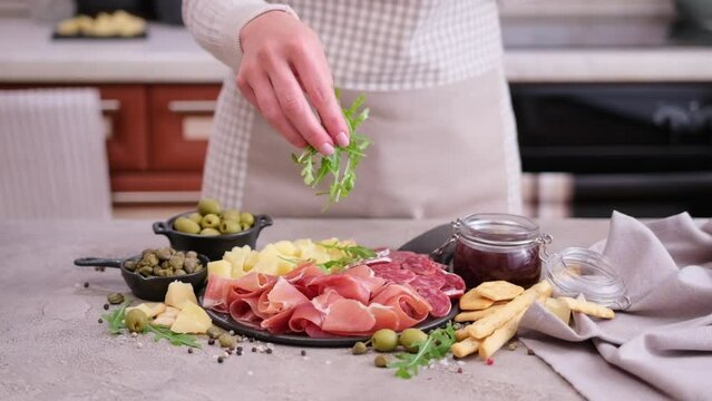 making meat and cheese antipasto plater - woman pouring arugula to serving board with cheese, prosciutto and fuet sausages