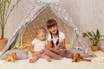 Indoor shot of dark haired female child with braids and her infant sister sitting on floor near wigwam and using cell phone for watching cartoons or interesting videos, playing in peetee together. © sementsova321