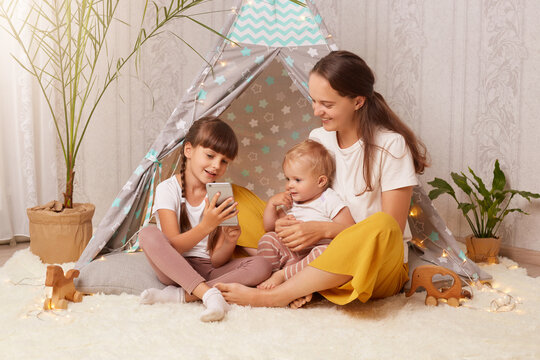 Playful caucasian woman and female children using modern smartphone for watching cartoon or photos in wigwam, mother and daughters playing together during free time at home. - Powered by Adobe