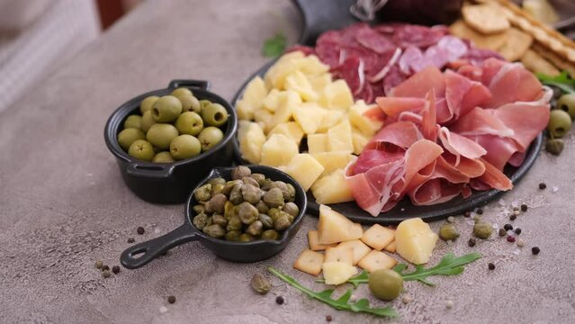 making meat and cheese antipasto plater - woman adding capers and olives to serving board with cheese, prosciutto and fuet sausages