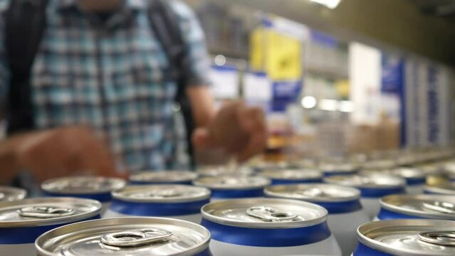 Close-up of many aluminium cans of beer or another drink on a store shelf and a man with a shopping trolley takes a couple