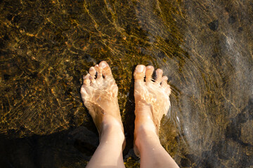 Two women's bare feet stand on a stone under shallow running water on a summer sunny day. Vacation and travel concept