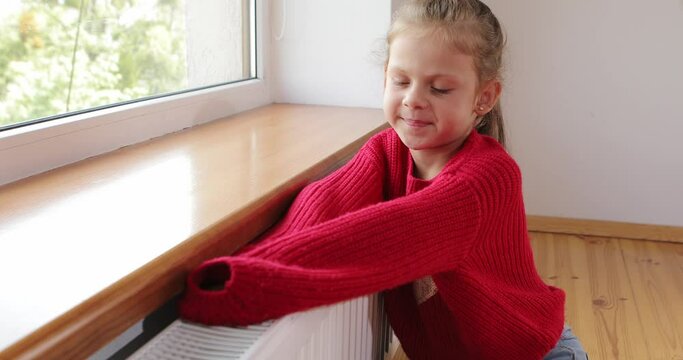 a little girl warms her hands and feet on a radiator