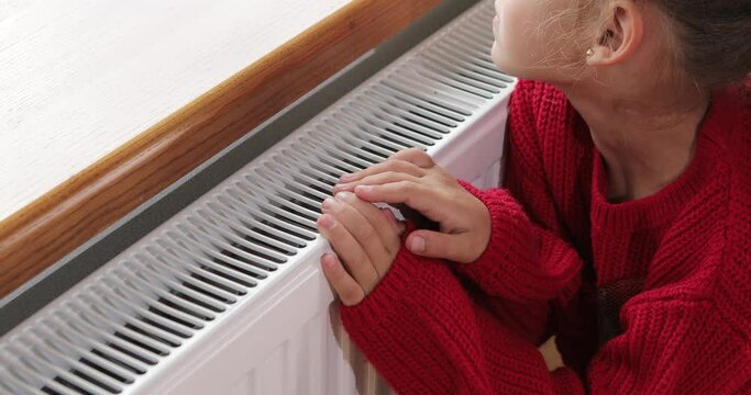 a little girl warms her hands and feet on a radiator