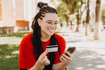 A cheerful young woman is making online shopping from her phone in park .