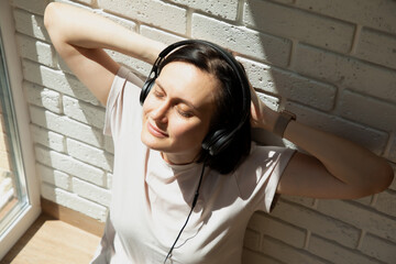 A woman near the window wearing black headphones and listening to music with her eyes closed. View...