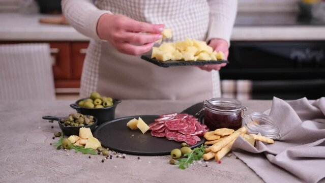 making meat and cheese antipasto plater - woman putting pieces of parmesan hard cheese on stone serving board at domestic kitchen