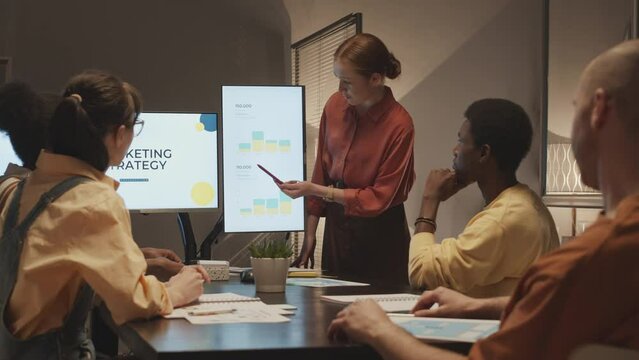 Medium slowmo of young Caucasian woman making presentation to colleagues from marketing agency, talking about successful promotion strategy and pointing at chart with numbers on projection screen