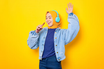 Dancing young Asian woman in jeans jacket wearing headphones for listening to music and singing via mobile phone isolated over yellow background