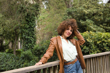 Naklejka na ściany i meble Mature and attractive woman, with curly brown hair, wearing a brown leather jacket, shirt and jeans, touching her hair in a sexy attitude. Concept fashion, trend, beauty, sensuality, curls, hairstyle.
