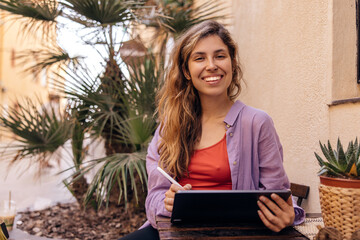 Happy young caucasian woman browsing online website on tablet sitting outdoors. Brown-haired girl smiles looking at camera, wears shirt. Technological device concept.