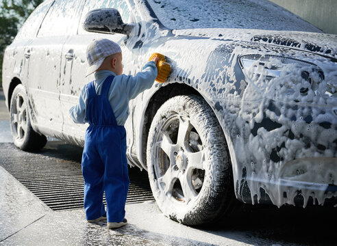 Toddler boy learning to wash car, wiping with sponge soapy foam on side of vehicle. Child getting new experience of cleaning automobile. Small kid cleaner soaping car by hand with sponge outdoors.
