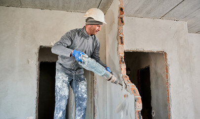 Man builder in construction helmet drilling wall with hammer drill. Male worker using drill breaker...