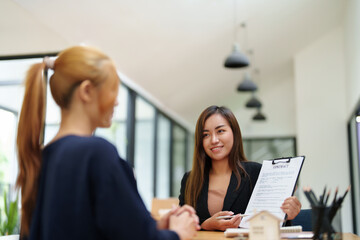 Portrait of an Asian female bank employee asking a customer to read the contract before signing to...