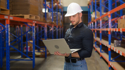 Young caucasian male worker wearing helmet using laptop in modern warehouse.