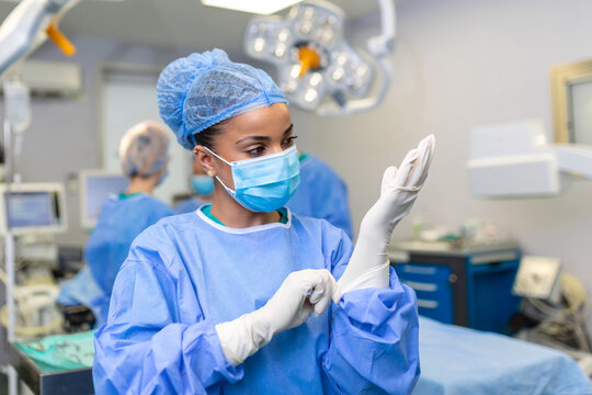 Young Female Doctor Prepares For Surgery, Wears Blue Surgical Gloves, In A Coat And Mask