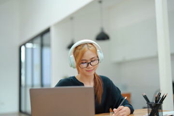 Portrait of a young Asian woman sitting at work wearing headphones over her ears to listen to music...