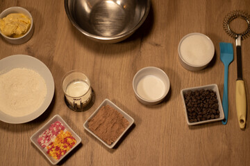 ingredients to make a chocolate sponge cake with cocoa nibs, sugar powder, chocolate powder, butter, flour, milk, yeast, sugar, with hearts and coloured stars, white, pink and orange on a wooden table