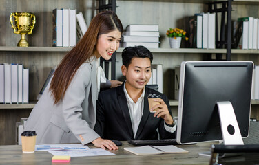 Asian cheerful professional successful businesswoman manager entrepreneur mentor in formal suit standing helping explaining discussing with young businessman employee sitting working with computer